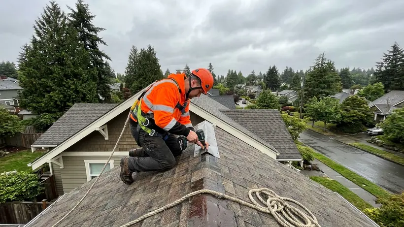 A roofing professional wearing a safety harness installing shingles on a steep roof.